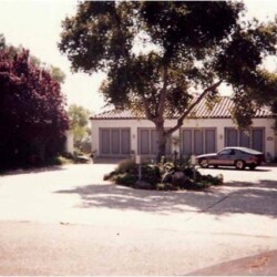 A large house with closed shutters and a parked car in front, surrounded by trees and shrubs on a sunny day.