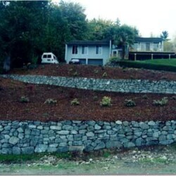 A house sits on a hill with terraced stone retaining walls and young plants in landscaped beds.
