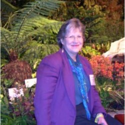 Woman in a purple blazer and blue scarf sitting in front of lush plants and flowers, smiling at the camera.